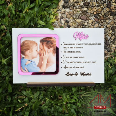 Placa decorativa branca com foto de mãe e filha e texto em português, moldura rosa, sobre erva e pedra.