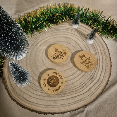 Três porta-copos de madeira com mensagens de Natal sobre um tronco de madeira rodeado por decoração natalícia.