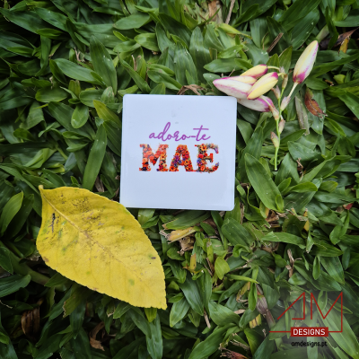 Caixa branca com texto 'adoro-te MÃE' sobre folhas verdes, folha amarela e flores brancas ao lado, logo DM DESIGNS vermelho