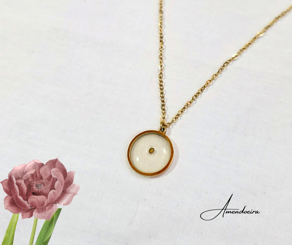 Colar da Fé medalha - semente de mostarda Colar dourado com pendente redondo de vidro com ponto dourado, sobre fundo branco com flor rosa e texto Amendoeira