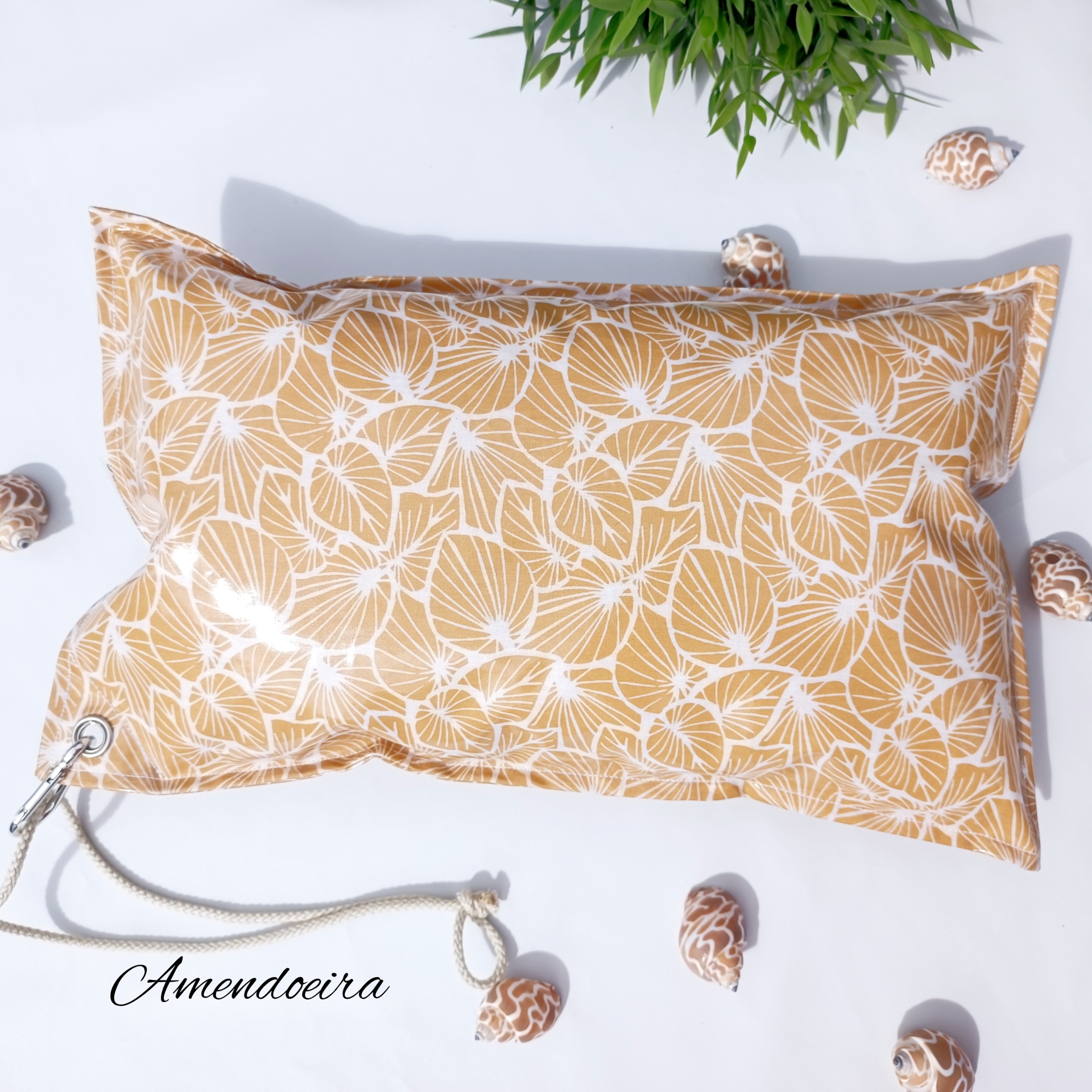 Almofada Mustard plants Almofada retangular bege com padrão branco e corda cinza, conchas e planta verde ao redor