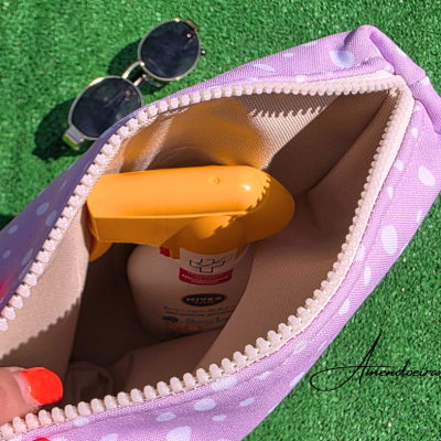 Necessaire roxa com bolinhas brancas aberta com frasco de spray laranja e óculos de sol pretos numa superfície verde.