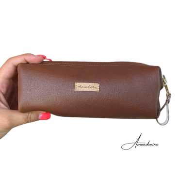 Estojo castanho tipo pele com etiqueta 'Amendoeira' e fecho de correr vermelho na mão.