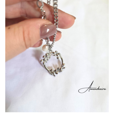 Colar prateado com pendente em forma de raminho e pérola segurado por mão com unhas castanhas, fundo branco com texto Amendoeira