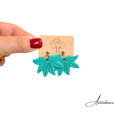 Brincos verdes em forma de folhas presos a um cartão bege, segurados por uma mão.