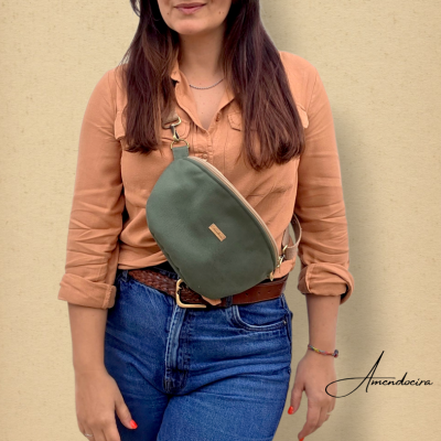 Mulher com mala pochete verde ao peito, camisa castanha e jeans azuis, fundo bege
