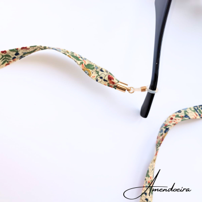 Cordão para óculos floral com metal dourado ligado a óculos preto sobre fundo branco