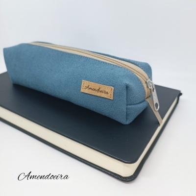 Estojo azul com fecho bege sobre caderno preto, fundo branco.