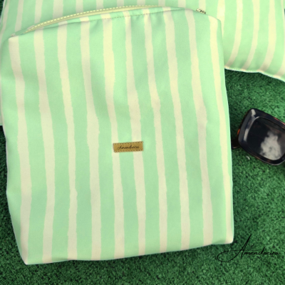 Estojo verde claro com riscas brancas e etiqueta Amandine, ao lado de óculos escuros, sobre relva artificial.