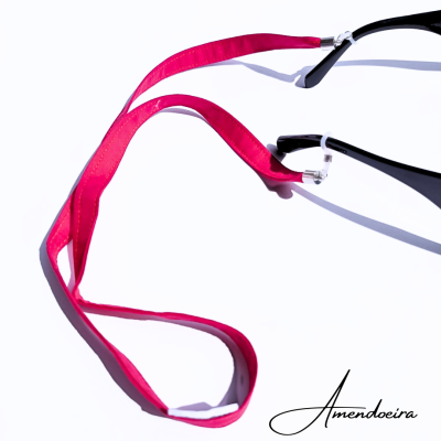 Cordão para óculos rosa com presilhas transparentes preso a óculos pretos sobre fundo branco.