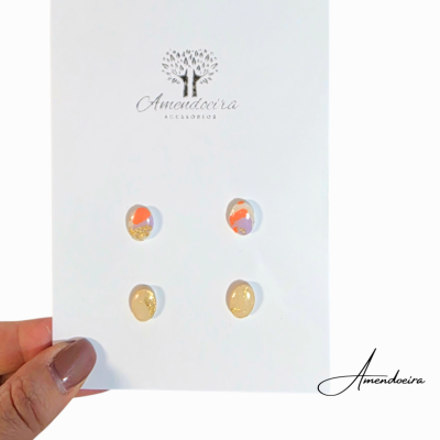 Cartão branco com brincos ovais coloridos e dourados da marca Amendoeira, segurado por mão feminina