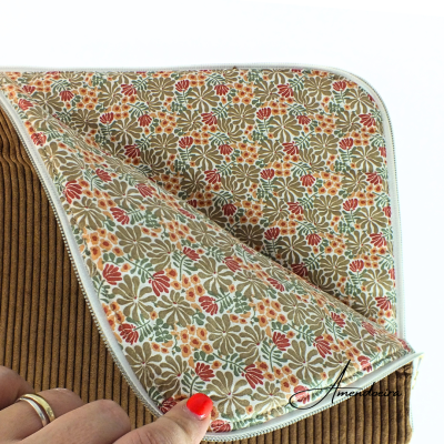 Manta floral com borda branca sobre tecido canelado castanho, segurada por uma mão.