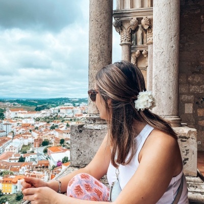 Mulher com blusa branca e calças estampadas numa varanda de pedra com vista urbana