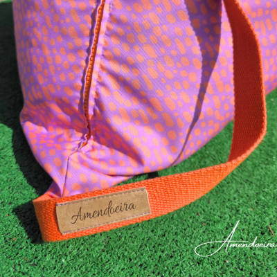 Bolsa rosa com padrão lilás, alça laranja e etiqueta 'Amendoeira' sobre relva verde