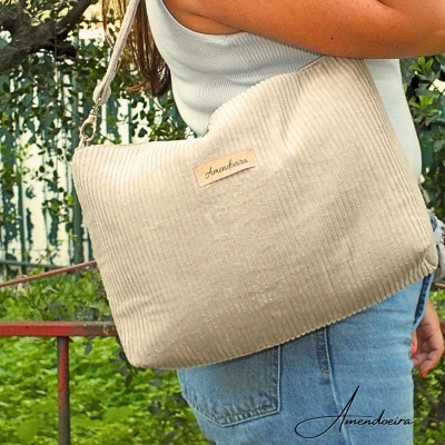 Mala de ombro bege claro com etiqueta Amendoeira, usada por pessoa com top branco e jeans, cenário ao ar livre