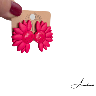 Brincos flor vermelhos em cartão castanho, segurados por mão com unhas vermelhas.