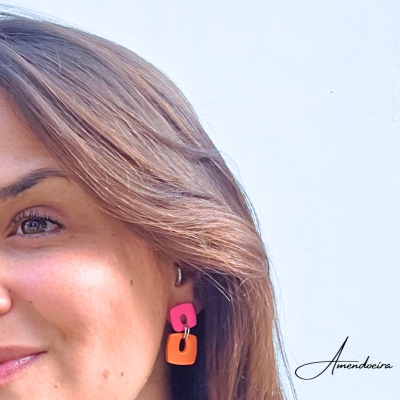 Mulher com brinco quadrado rosa e laranja, fundo branco com palavra Amendoeira