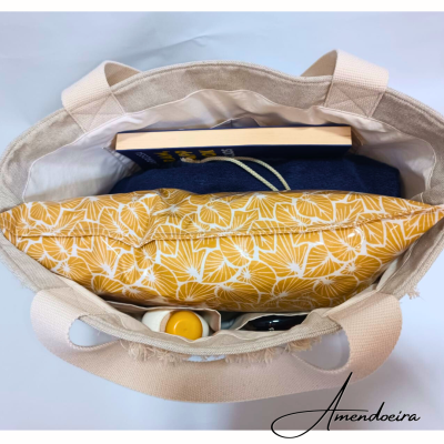 Saco de tecido cru com almofada amarela, livro azul, roupa azul e garrafa visíveis de cima