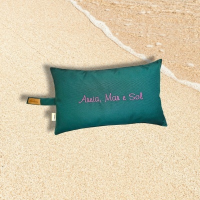 Almofada verde escura com texto bordado na areia junto a uma onda do mar