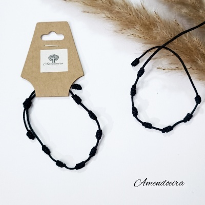 Pulseiras pretas com nós em cordão sobre fundo branco com embalagem e decoração de ervas secas