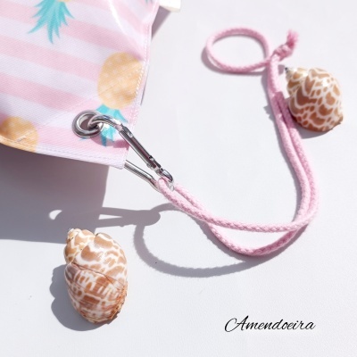 Saco rosa com padrão de abacaxis, argola metálica e cordão rosa, conchas naturais ao lado, texto 'Amendoeira'