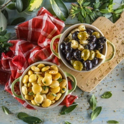 Duas taças com diferentes tipos de azeitonas em uma superfície azul com pano vermelho e folhas verdes