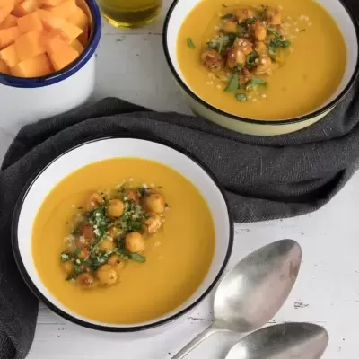 duas tigelas com sopa laranja cremosa e grão-de-bico, colher metal, azeite, pedaços de abóbora