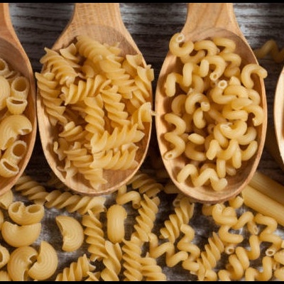 Quatro colheres de madeira com diferentes tipos de massa seca, sobre superfície de madeira