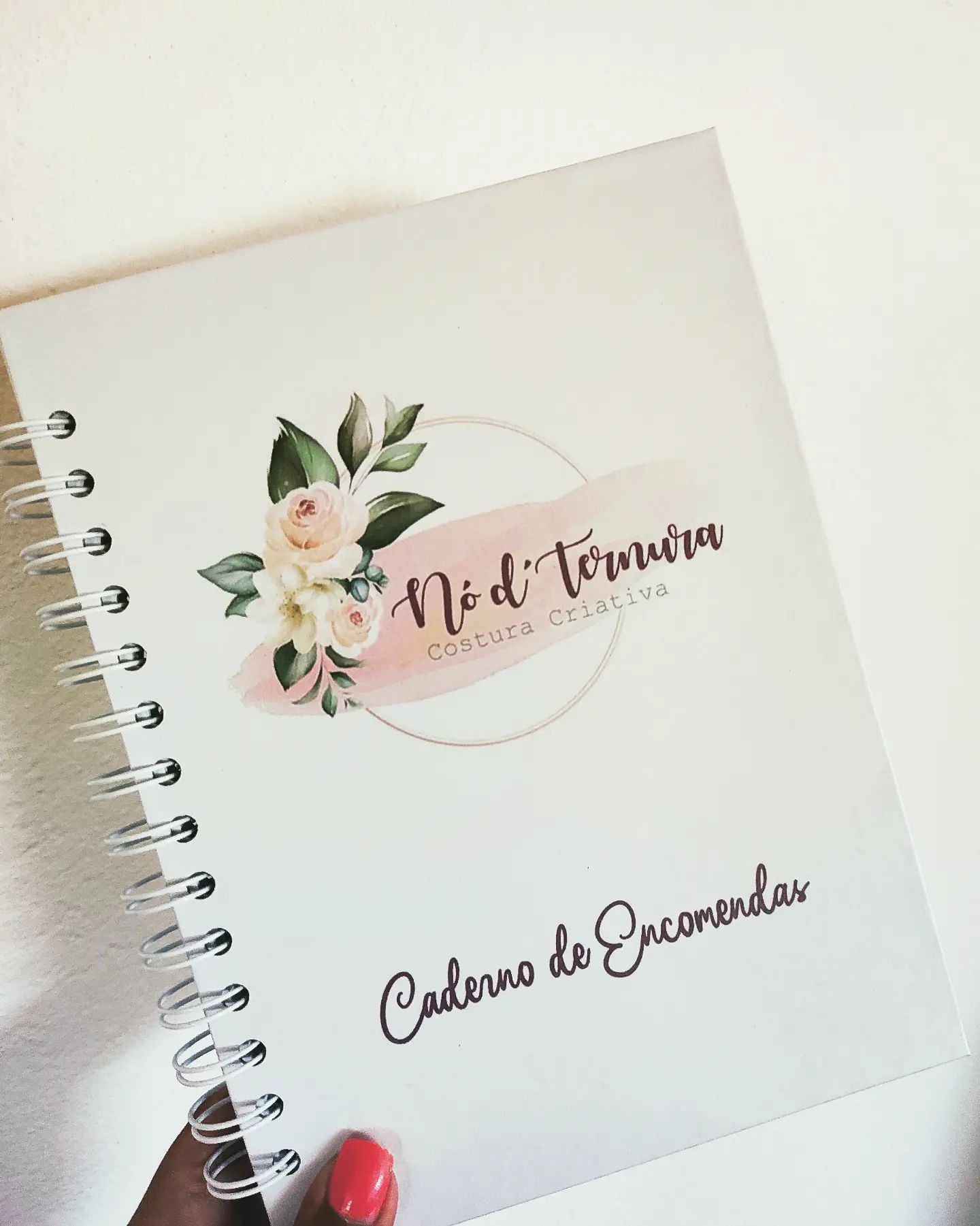 Caderno branco com capa decorada, texto e flores, encadernação espiral e mão segurando.