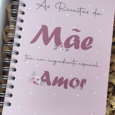 Caderno espiral rosa com texto 'As Receitas da Mãe têm um ingrediente especial... Amor' e decoração floral