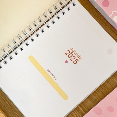 Agenda 2025 com capa branca em espiral sobre mesa de madeira com cadernos cor-de-rosa