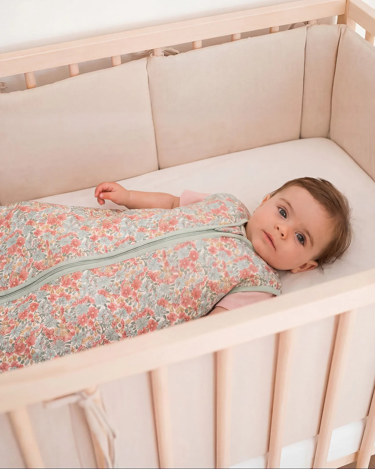 Bebé deitado no berço com saco de dormir floral colorido e proteção acolchoada bege.