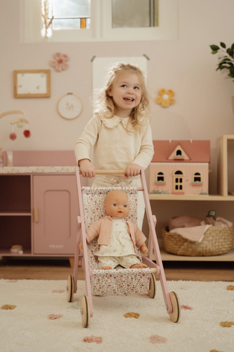 Criança loira a empurrar carrinho de boneca rosa com tecido floral num quarto infantil decorado