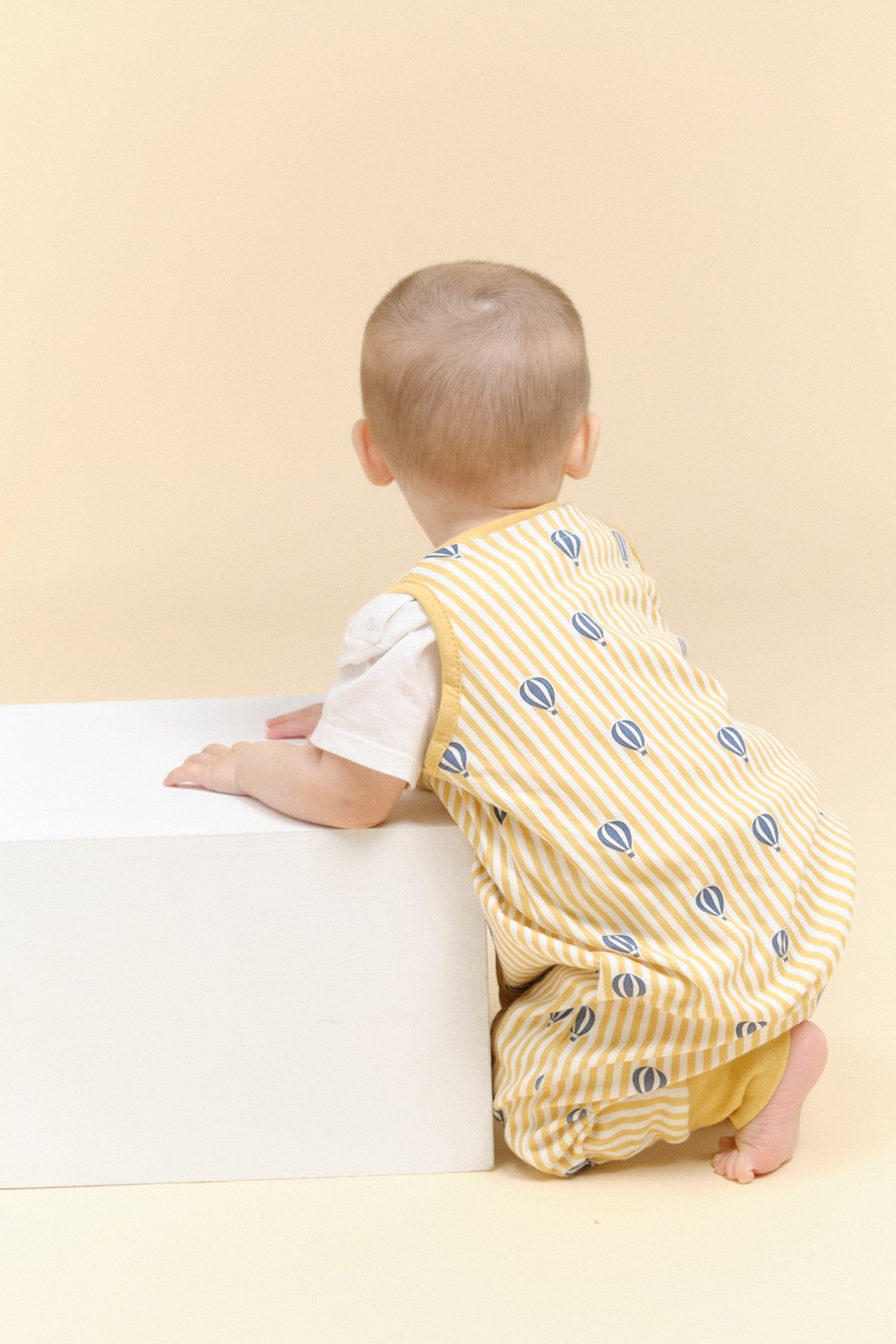 Bebé com roupa amarela e colete estampado de balões, visto de costas apoiado numa superfície branca
