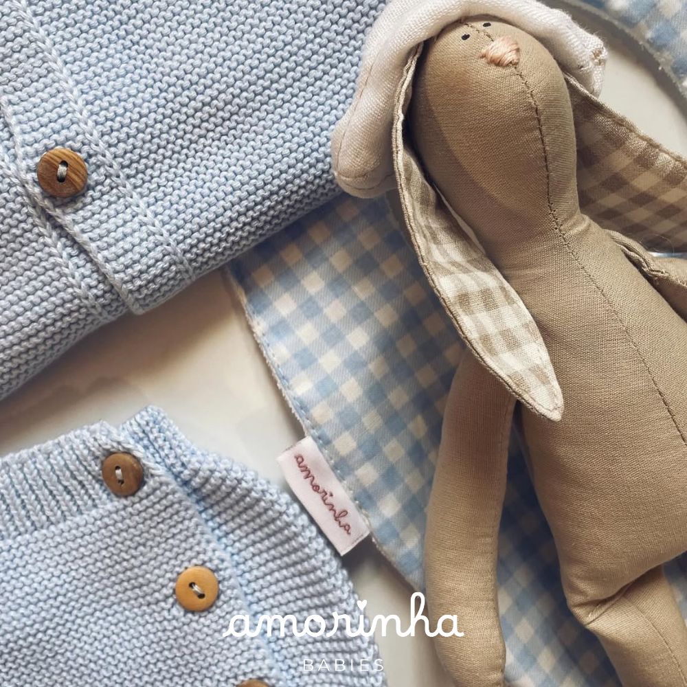 Roupa de bebé azul com botões de madeira, bandana xadrez azul e branca, e boneco de tecido bege com orelhas xadrez.