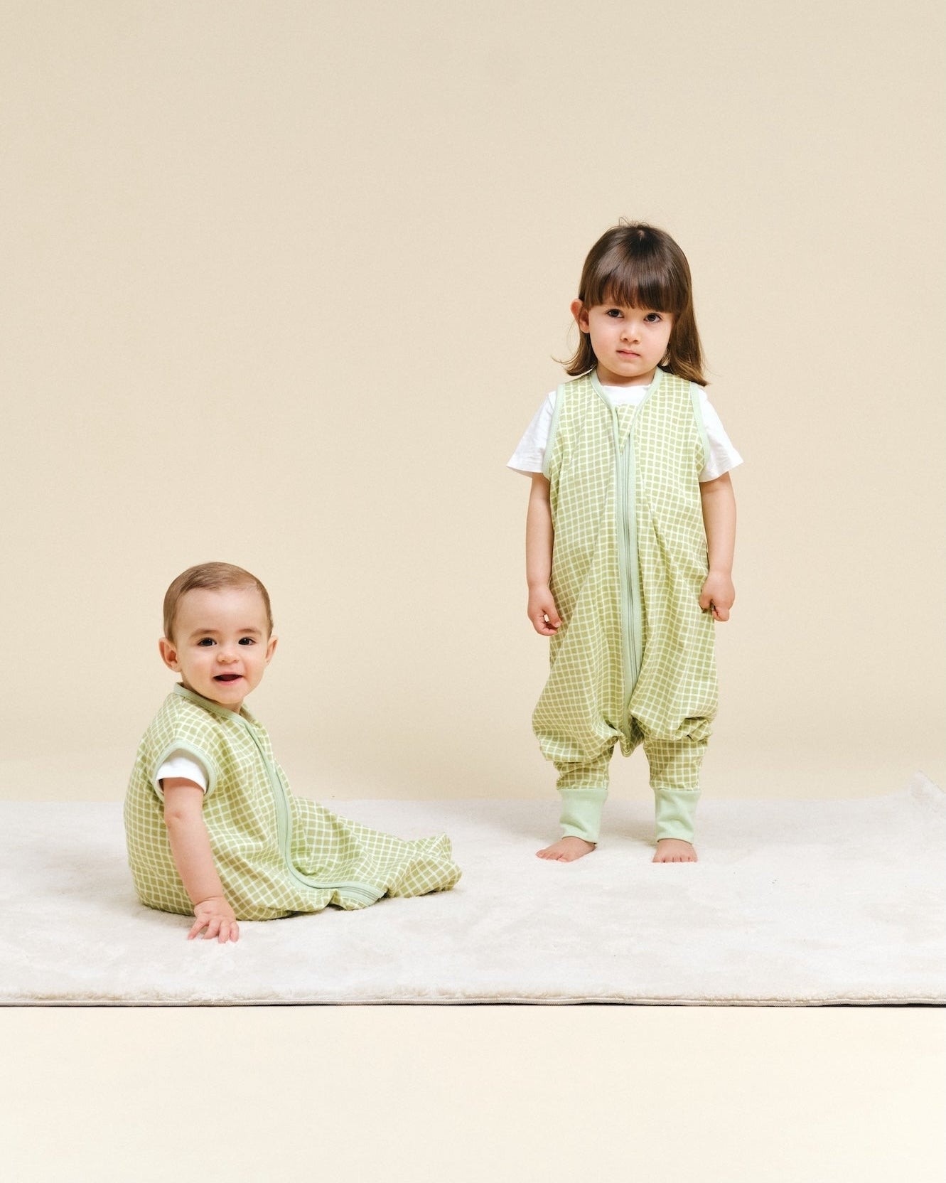 Dois bebés com roupa verde de padrão xadrez em fundo bege e tapete branco