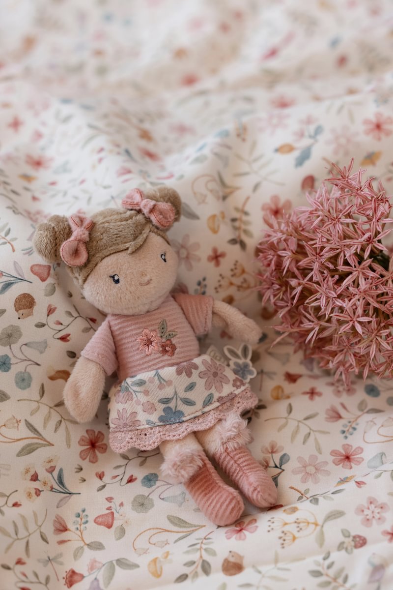 Boneca de peluche com roupa rosa e saia floral sobre tecido florido, ao lado de flores cor-de-rosa secas