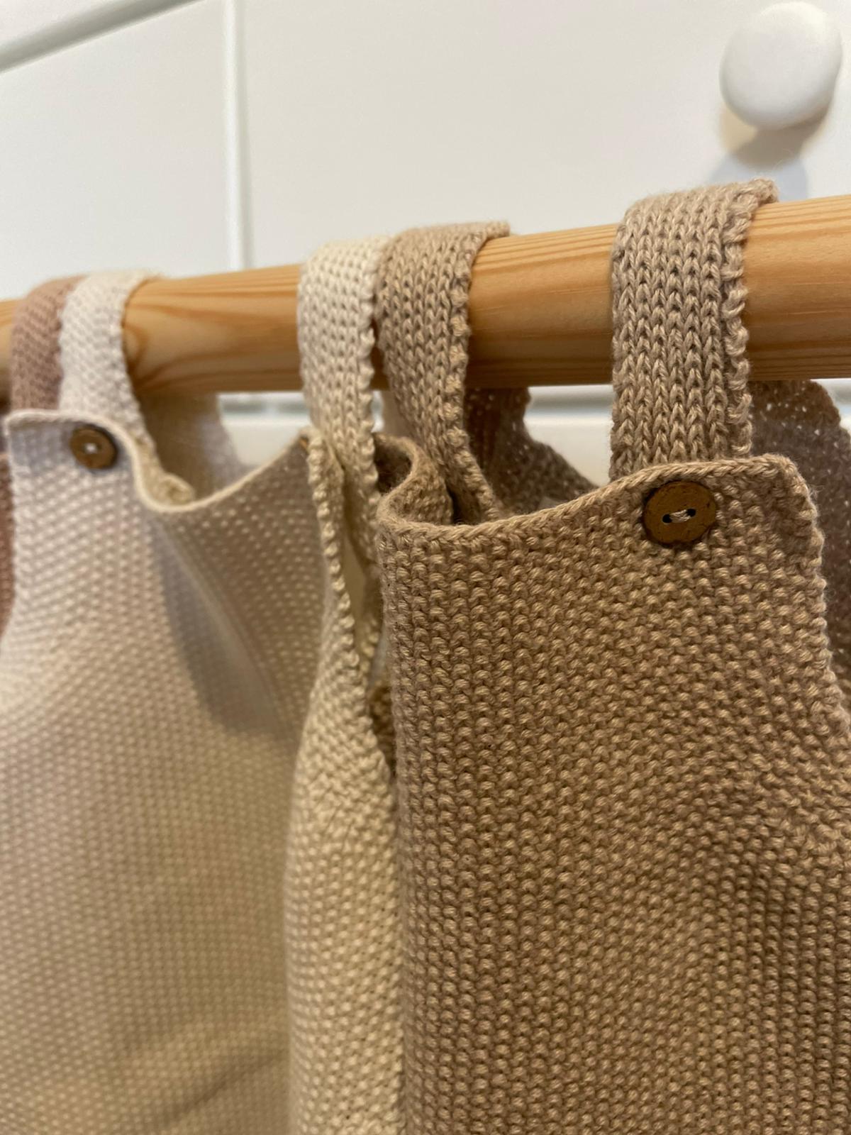 Peças de roupa tricotadas em tons naturais penduradas em varão de madeira.