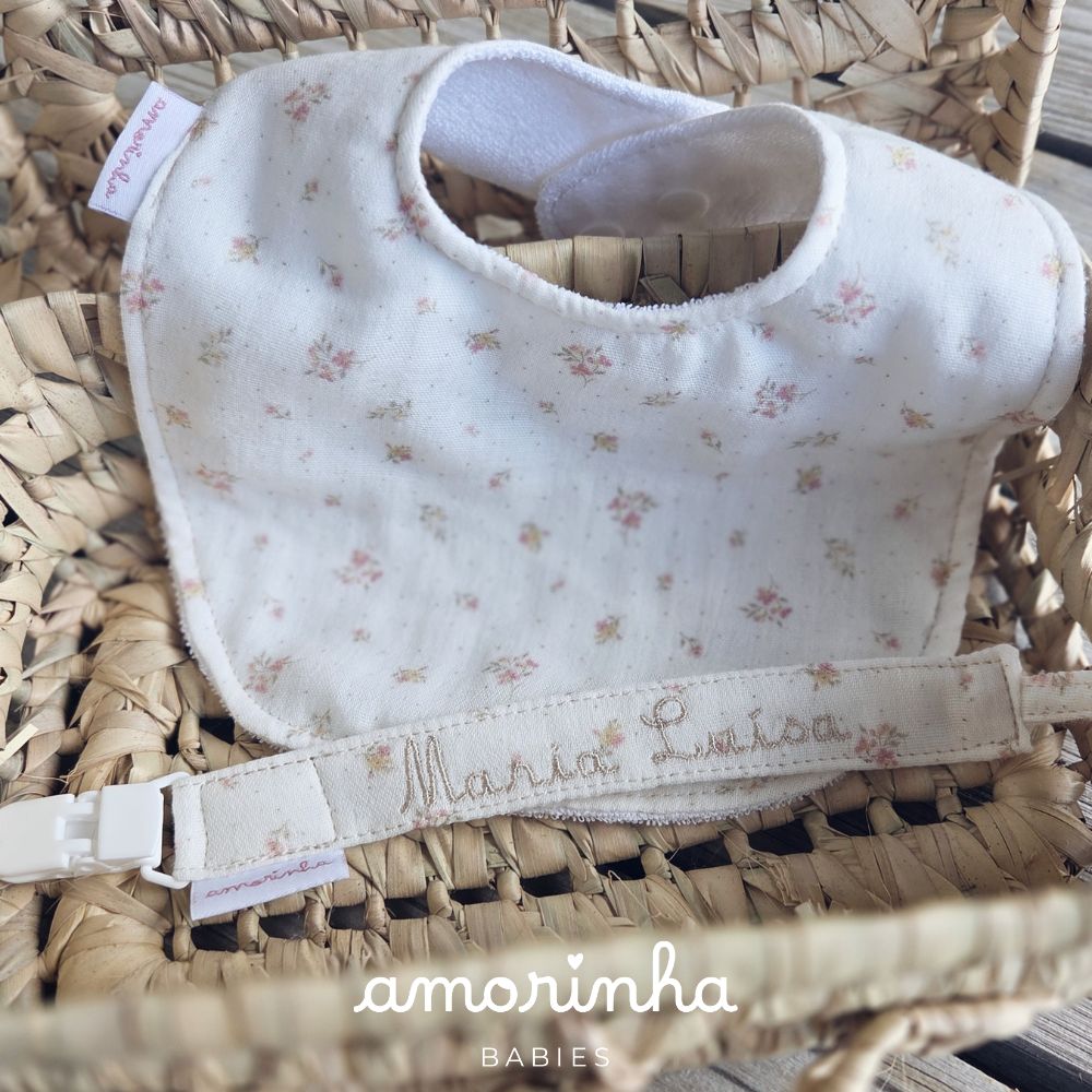 Babete e fita para chupeta com padrão floral e nome bordado numa cesta de palha