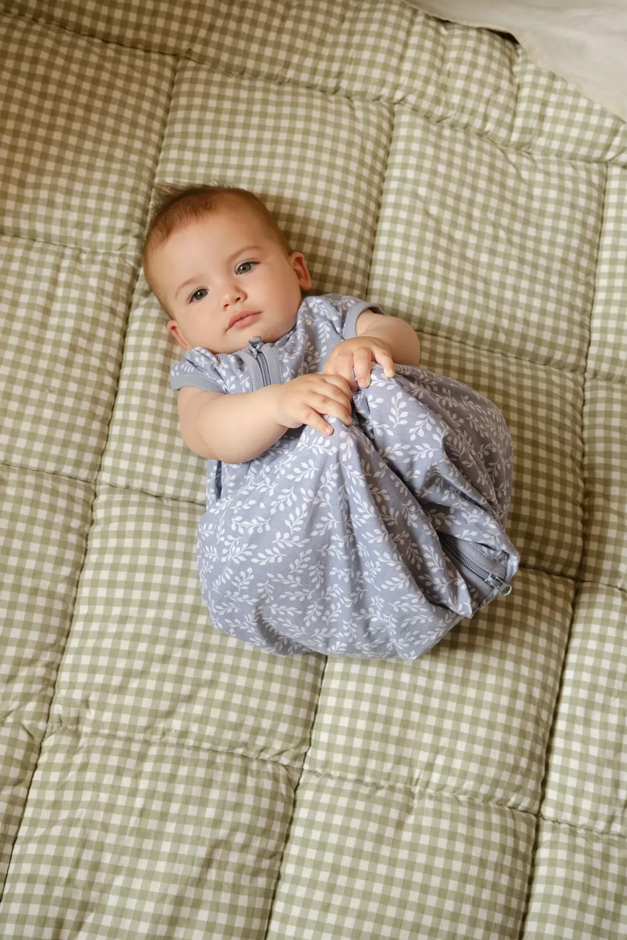 Bebé deitada num colchão acolchoado com padrão xadrez bege e branco, vestindo um saco de dormir azul claro com padrão branco de folhas.