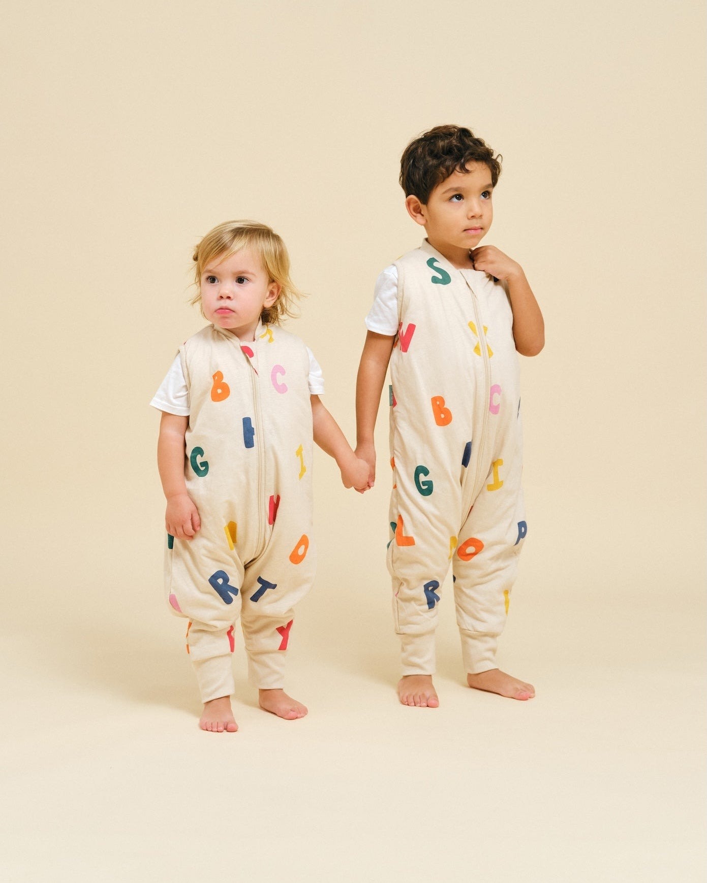 Duas crianças com babygrows creme com letras coloridas, em fundo creme.