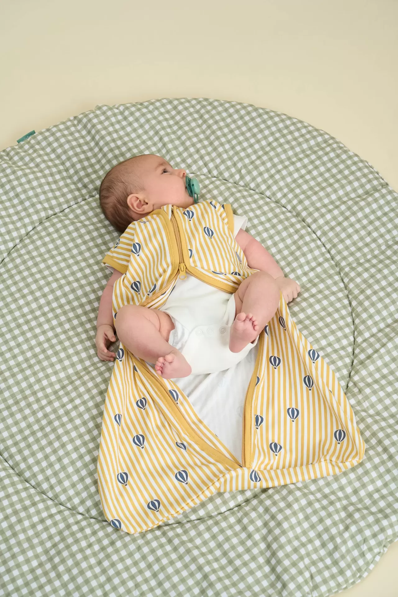 Bebé deitado num tapete acolchoado xadrez vestindo saco de dormir amarelo riscado com balões.