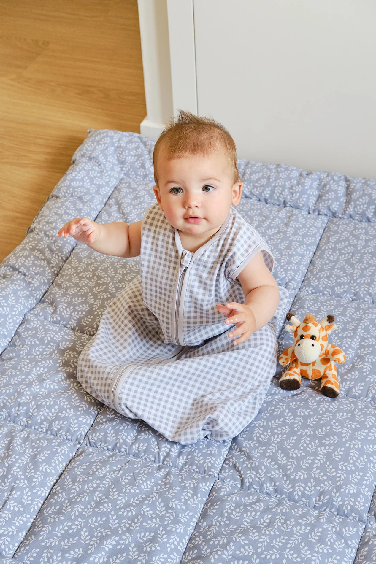 Bebé sentado num colchão almofadado azul claro padrão branco com saco de dormir cinzento e branco às quadrículas.