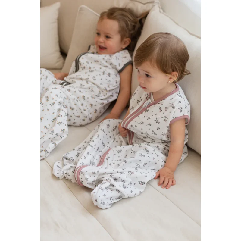 Dois bebés com sacos de dormir brancos de padrão floral sentados num sofá claro