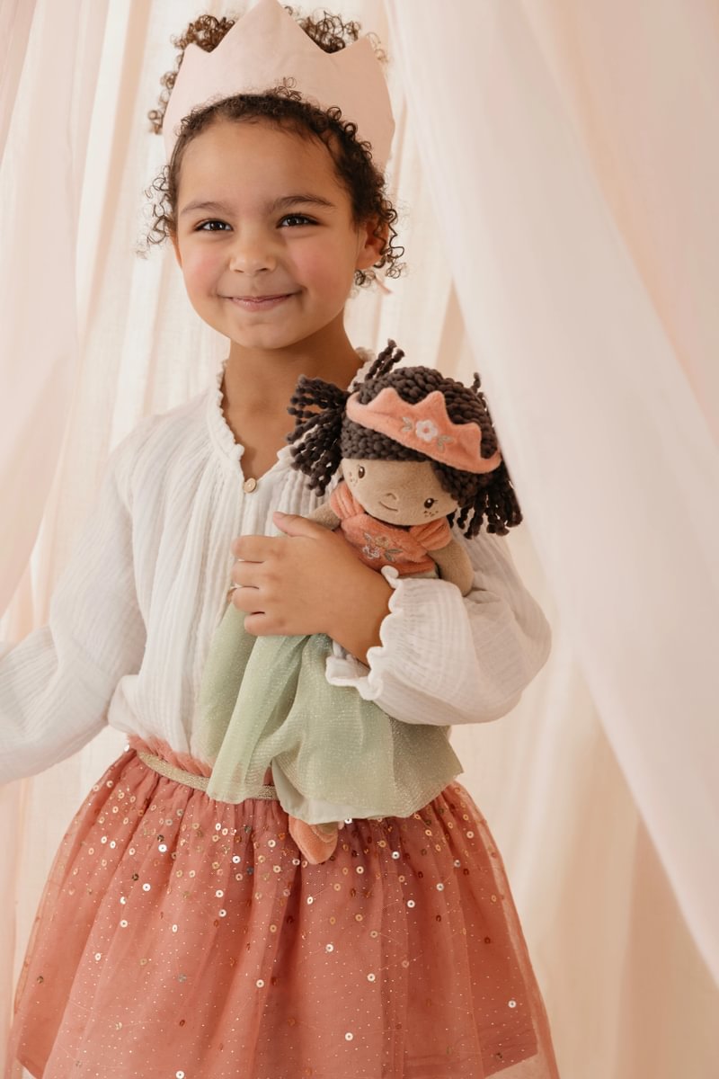 Criança com coroa rosa e vestido branco com saia rosa segurando boneca de pano vestida de verde.