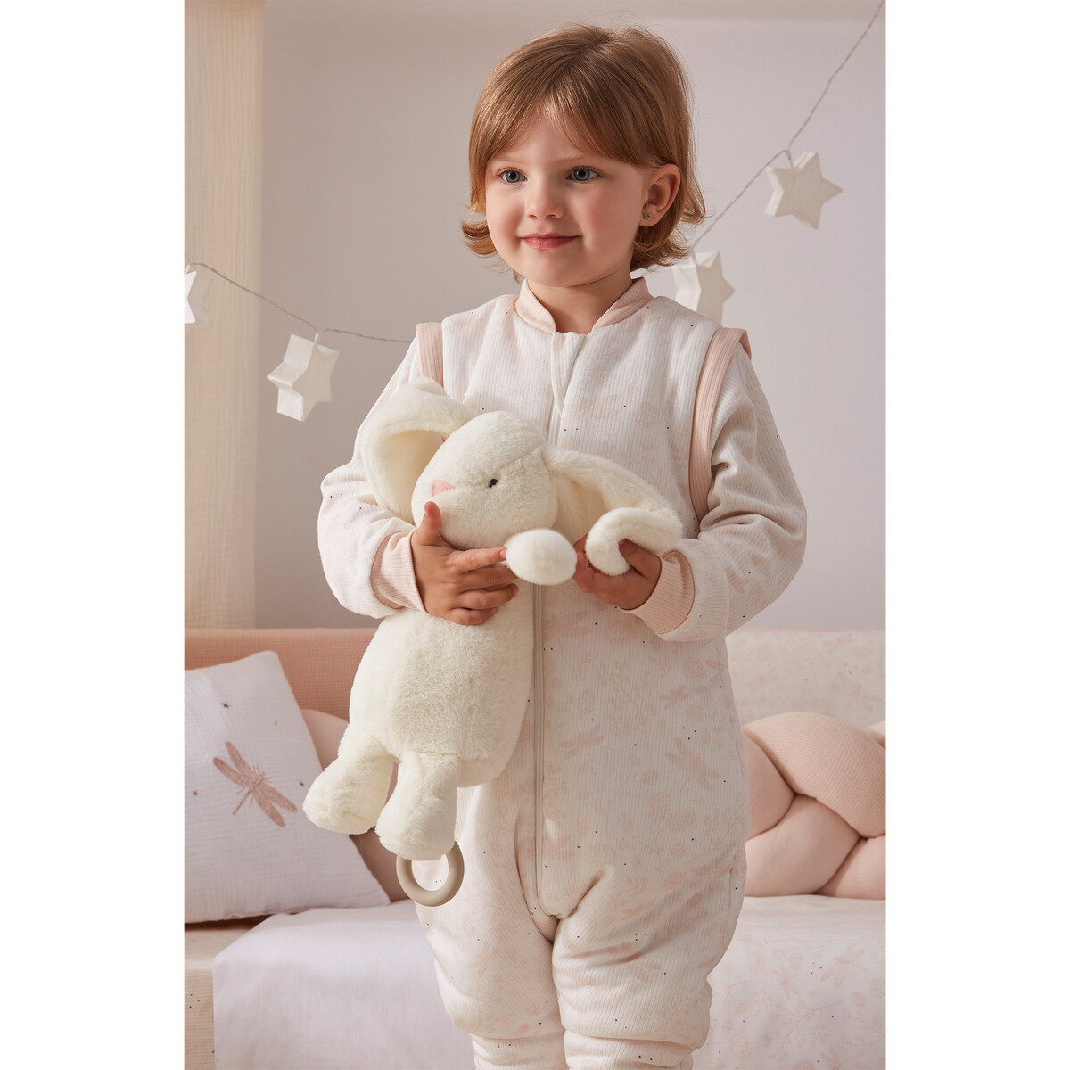 Menina com pijama rosa claro segurando peluche de coelho branco em quarto decorado com estrelas e almofada