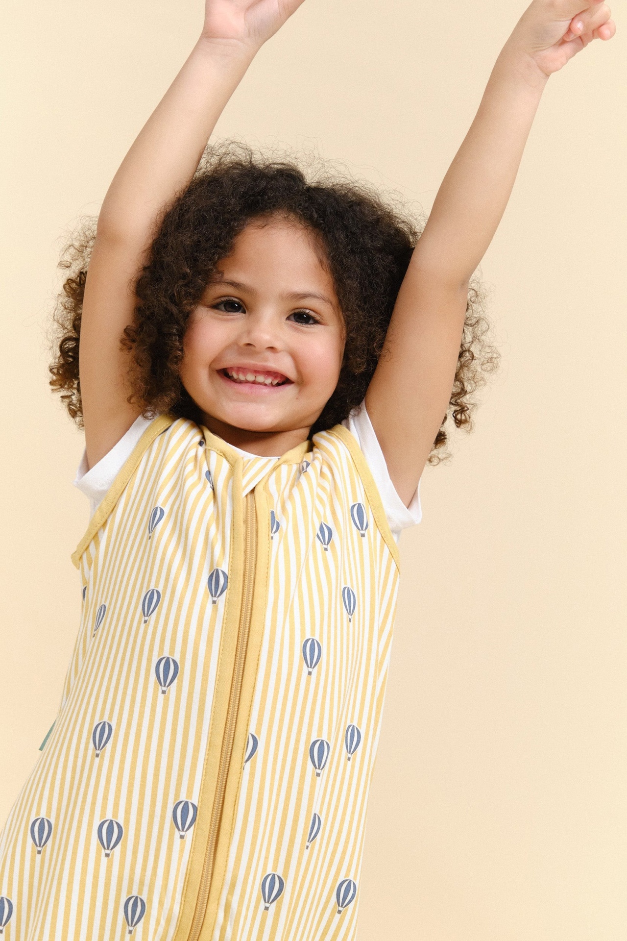 Criança sorridente vestindo saco de dormir amarelo com riscas e balões azuis.