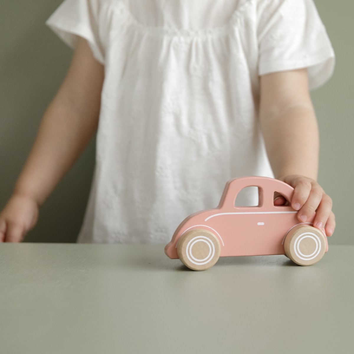 Criança com vestido branco a segurar carrinho de madeira rosa em superfície cinzenta