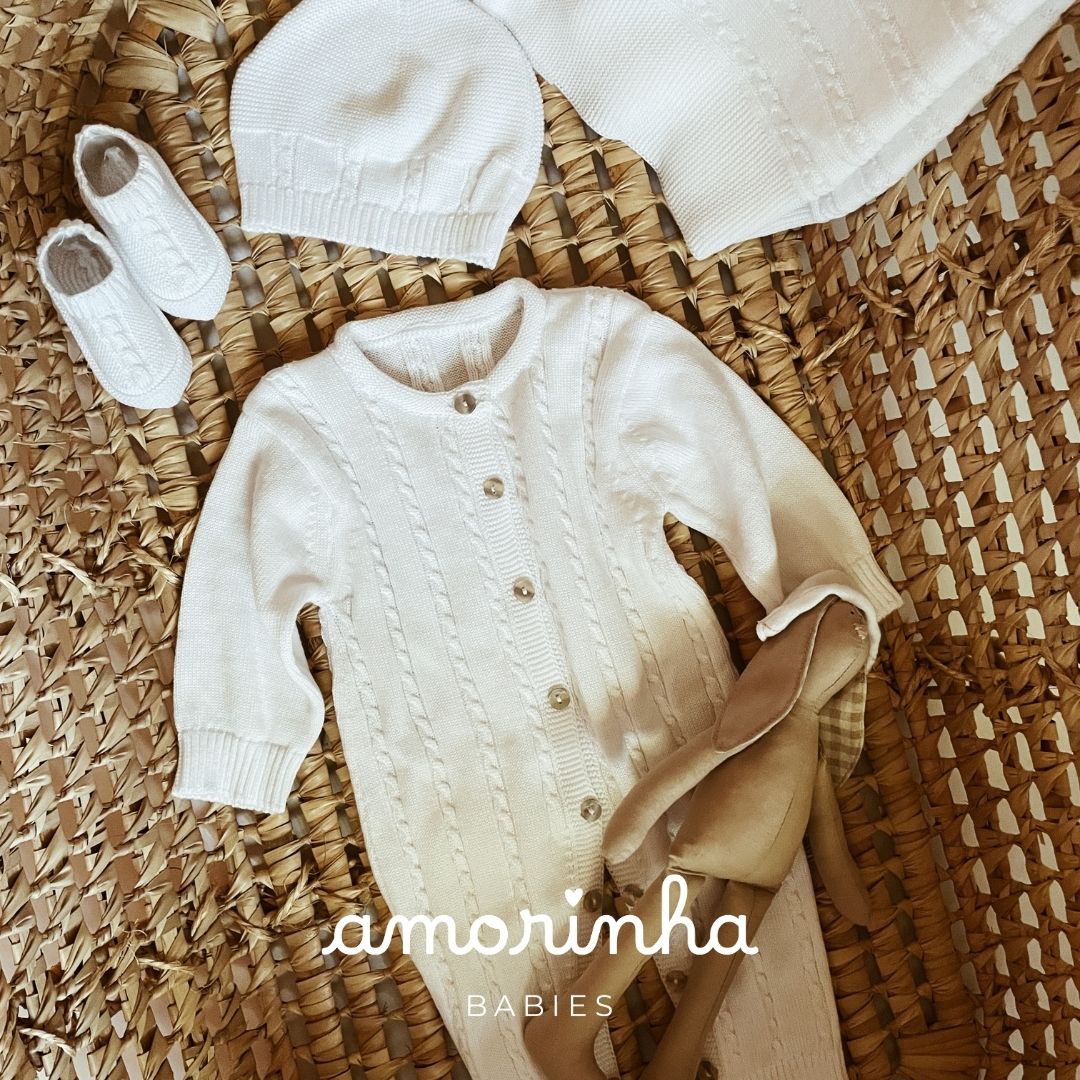 Conjunto de roupa de bebé branco com macacão, gorro e botinhas sobre tapete de fibras naturais.