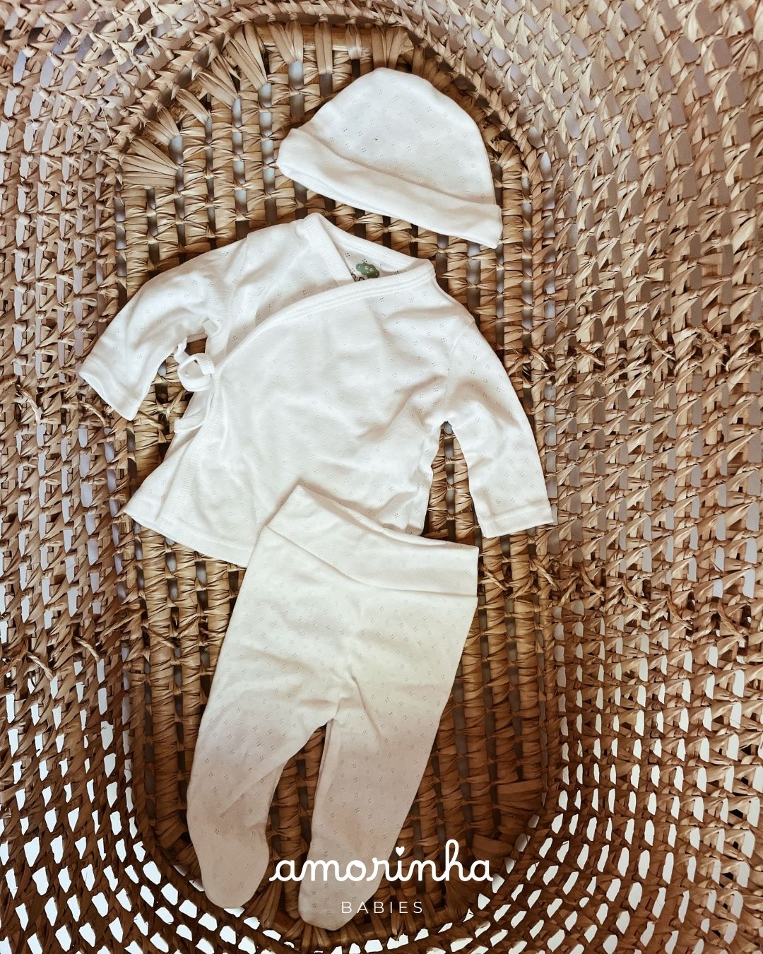 Conjunto de roupa branca para bebé com camisola, calças e gorro sobre tapete trançado